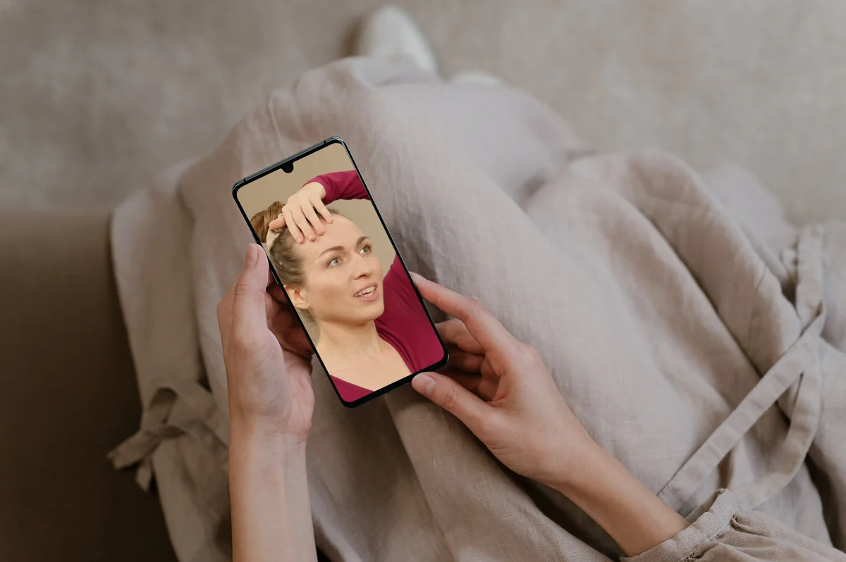 Hände halten ein Smartphone, auf dem Hanna Sacher eine Übung für die Stirn zeigt
